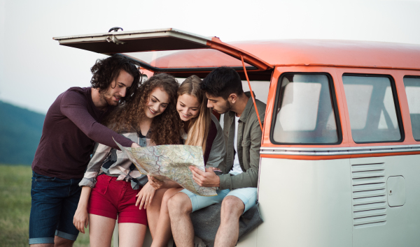 A group of cheerful young friends with a retro minivan on a roadtrip through countryside, looking at map.