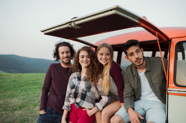 A portrait of group of young friends by a retro minivan on a roadtrip through countryside.