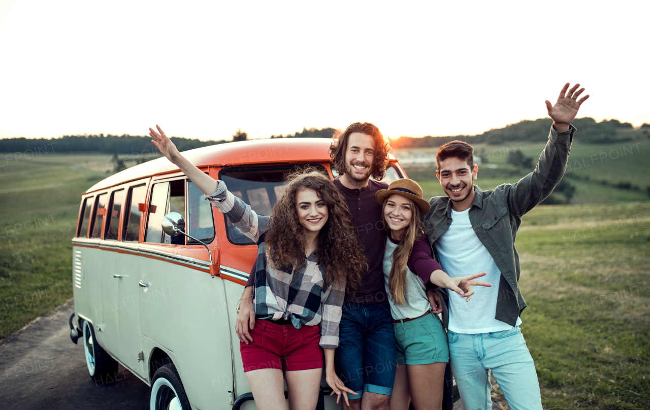 A group of young friends on a roadtrip through countryside, standing by a retro minivan at sunset.