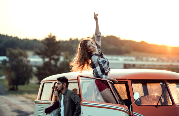 A group of young friends standing by a retro minivan on a roadtrip through countryside.