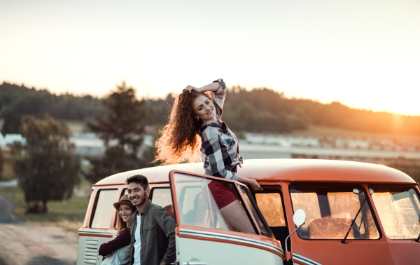 A group of young friends standing by a retro minivan on a roadtrip through countryside at sunset.