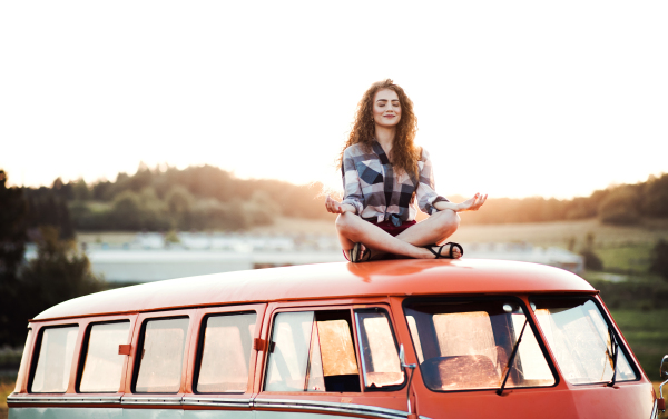 A young girl on a roadtrip through countryside, sitting on the roof of minivan doing yoga exercise.