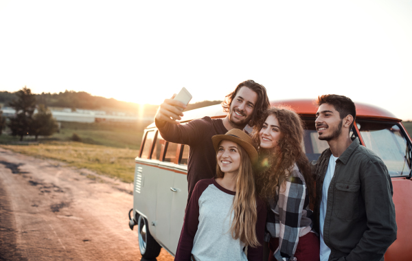 A group of cheerful young friends standing by a retro minivan on a roadtrip through countryside, taking selfie.