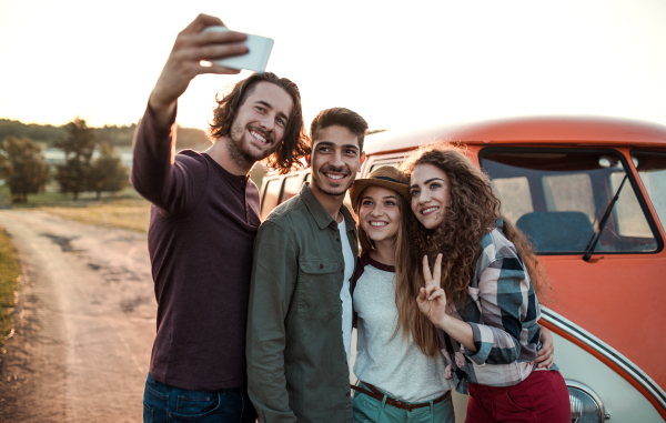 A group of cheerful young friends standing by a retro minivan on a roadtrip through countryside, taking selfie.