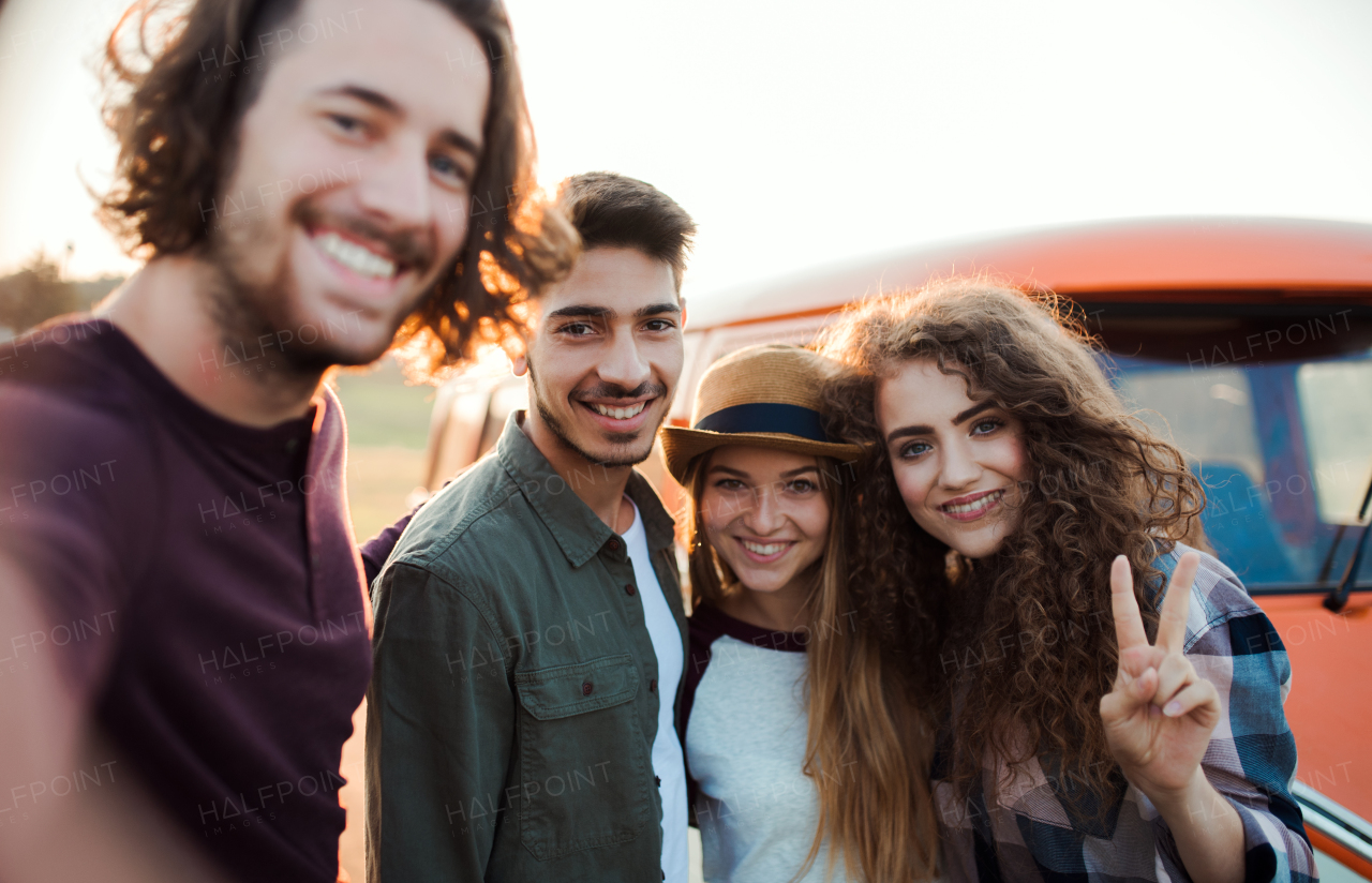 A group of cheerful young friends standing by a retro minivan on a roadtrip through countryside, taking selfie.