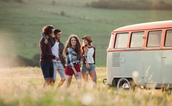 A group of young friends walking in front of a retro minivan on a roadtrip through countryside, walking arm in arm.