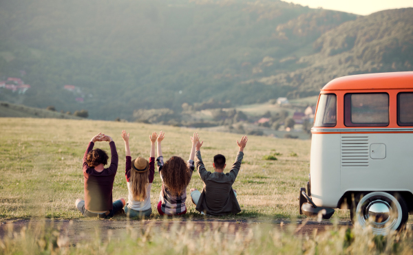 A rear view of group of young friends sitting on grass on a roadtrip through countryside, arms stretched.