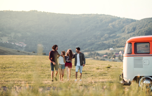 A group of young friends by a retro minivan on a roadtrip through countryside, walking arm in arm.