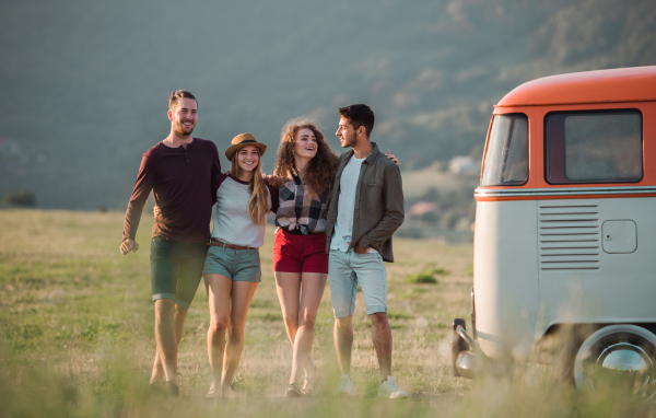 A group of young friends walking in front of a retro minivan on a roadtrip through countryside, walking arm in arm.