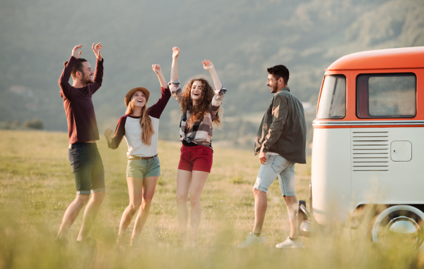 A group of cheerful young friends standing by a retro minivan on a roadtrip through countryside.