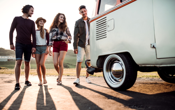 A group of cheerful young friends walking by a retro minivan on a roadtrip through countryside.