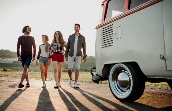 A portrait of group of young friends walking by a retro minivan on a roadtrip through countryside.