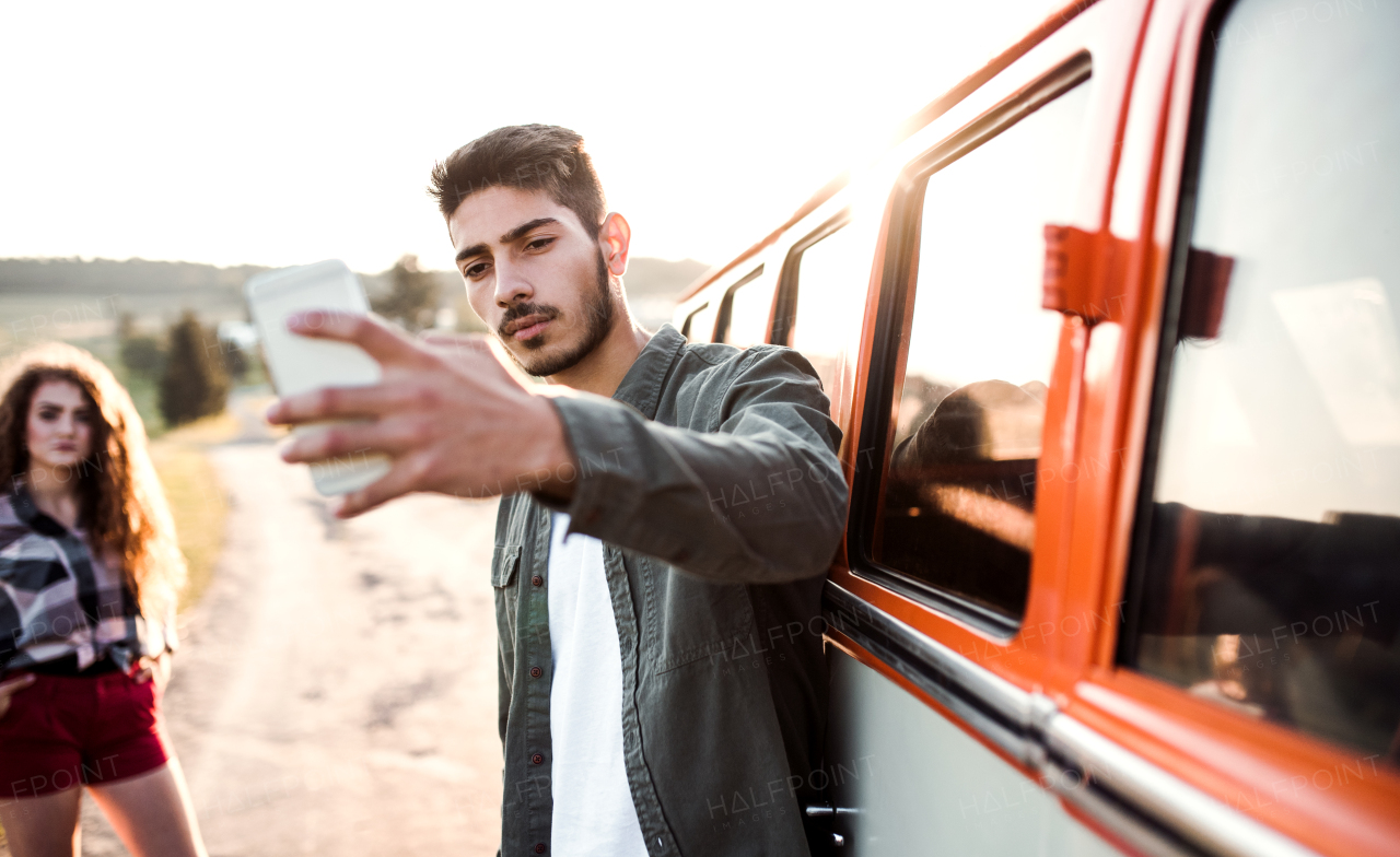 A young confident couple with smartphone by a car on a roadtrip through countryside, taking selfie.