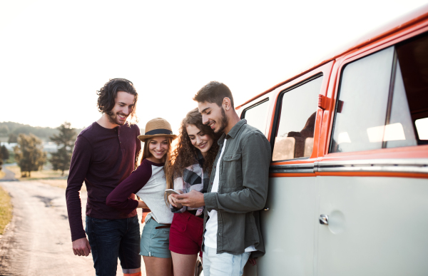 A group of cheerful young friends with a retro minivan on a roadtrip through countryside, using smartphone.