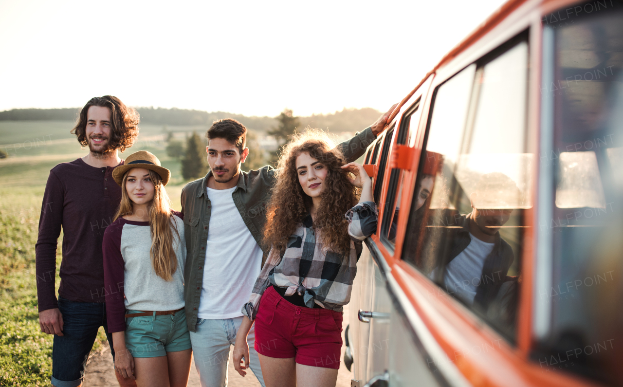 A portrait of group of young friends standing by a retro minivan on a roadtrip through countryside.