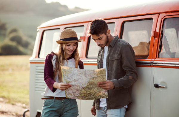 A young couple on a roadtrip through countryside, standing by retro minivan and looking at a map.