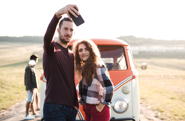 A group of young friends standing by a retro minivan on a roadtrip through countryside, taking selfie with smartphone.