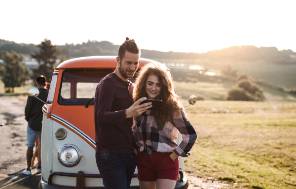 A young couple on a roadtrip through countryside standing by a minivan, using map on smartphone.