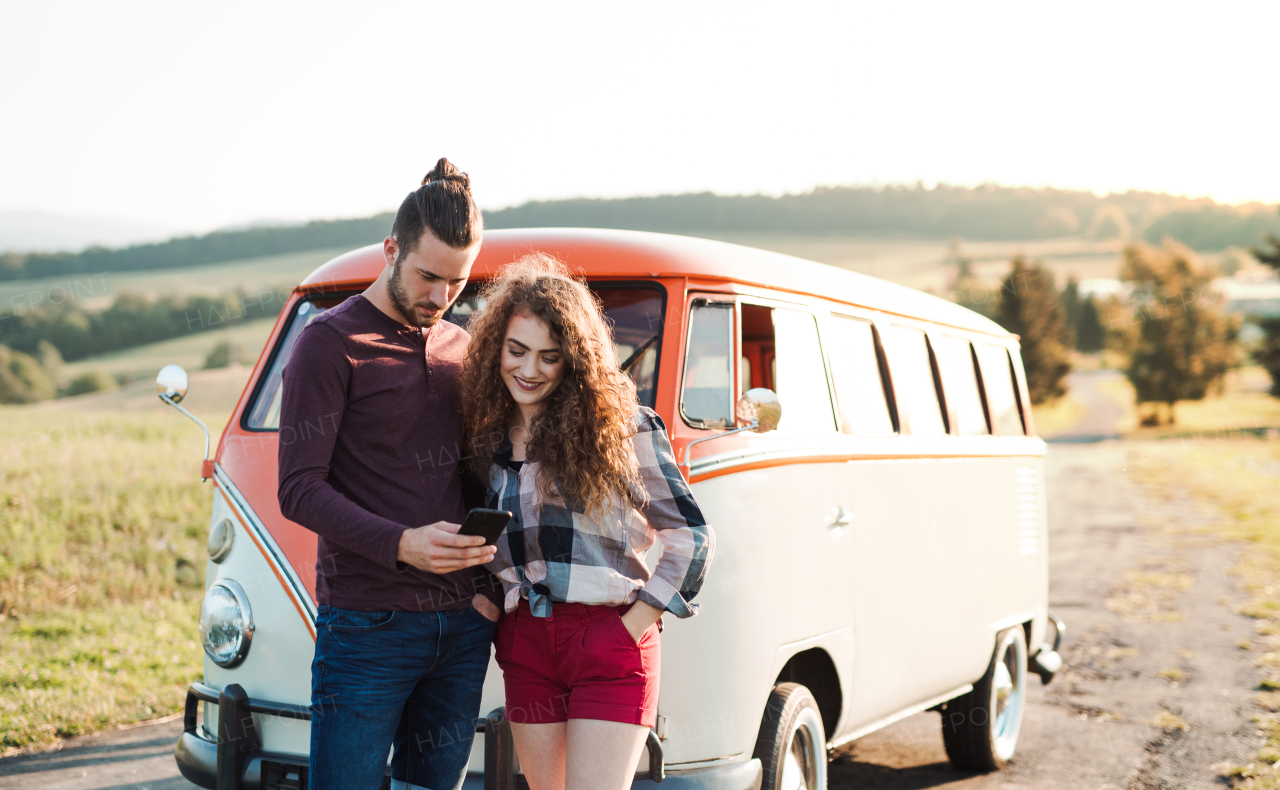 A young couple on a roadtrip through countryside standing by a minivan, using map on smartphone.