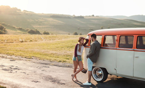 A young couple on a roadtrip through countryside, standing by retro minivan, talking.