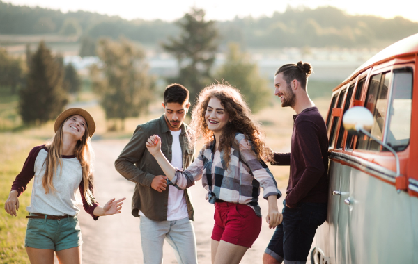 A group of cheerful young friends standing by a retro minivan on a roadtrip through countryside, dancing.