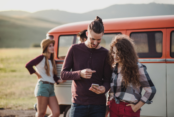 A young couple on a roadtrip through countryside standing by a minivan, using map on smartphone.