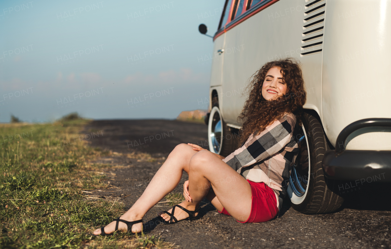 A young girl on a roadtrip through countryside, sitting on the ground by a retro minivan, eyes closed.