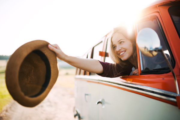 A cheerful young girl looking out of a car on a roadtrip through countryside, holding hat.