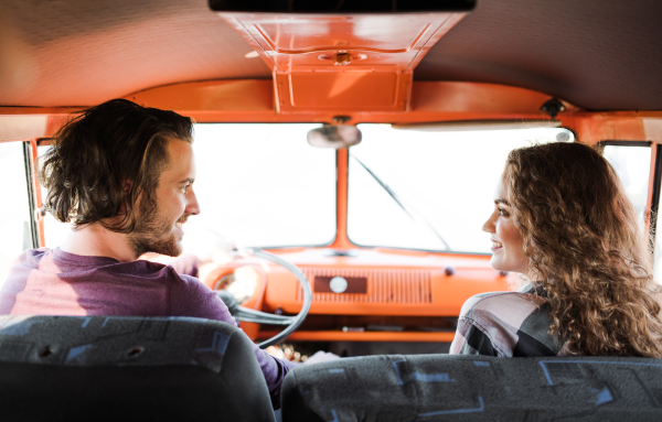 A rear view of young couple on a roadtrip through countryside, driving retro minivan.