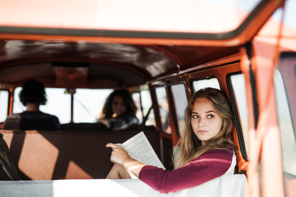 A group of cheerful young friends with a retro minivan on a roadtrip through countryside, one girl reading a book.