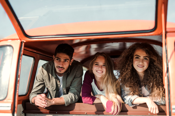 A group of cheerful young friends with a retro minivan on a roadtrip through countryside, looking out of window.