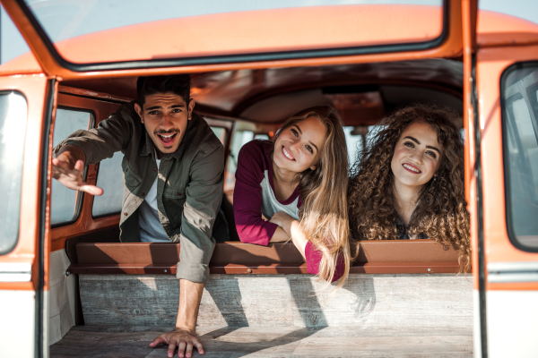 A group of cheerful young friends with a retro minivan on a roadtrip through countryside, looking out of window.