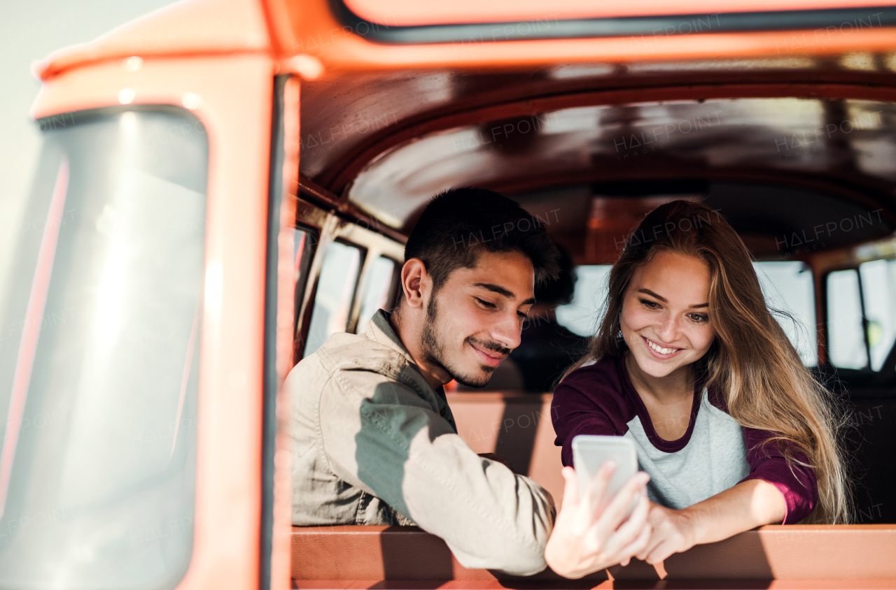 A group of young friends standing by a retro minivan on a roadtrip through countryside, taking selfie with smartphone.