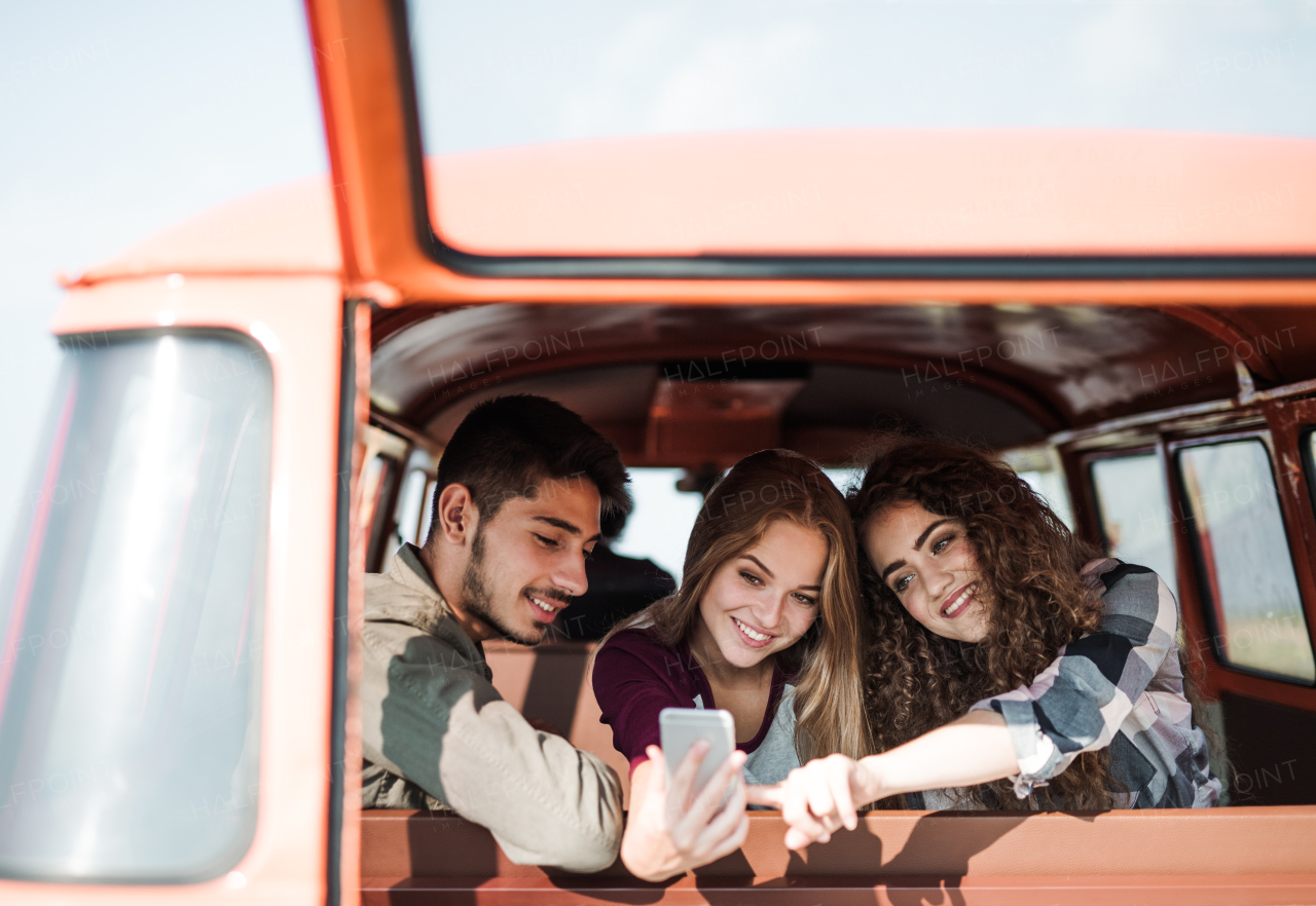 A group of cheerful young friends with a retro minivan on a roadtrip through countryside, taking selfie with smartphone.