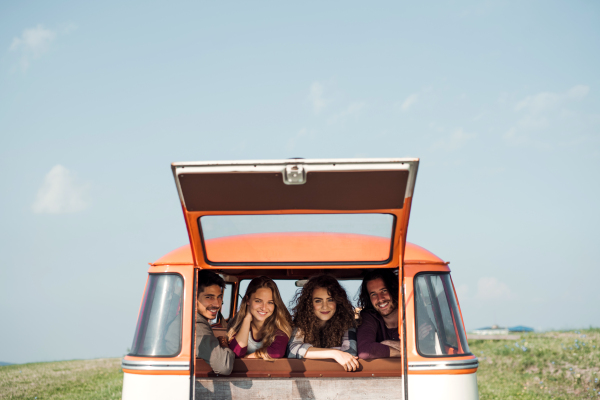 A group of cheerful young friends with a retro minivan on a roadtrip through countryside, looking out of window.