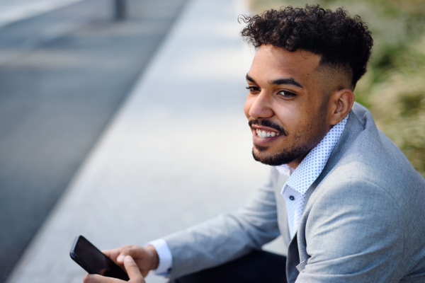 A portrait of young man sitting outdoors in city, using smartphone.