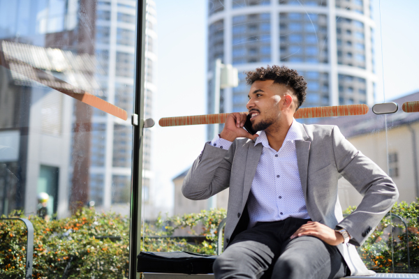 A portrait of young man student sitting outdoors on bus stop in city, using smartphone.