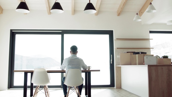 A rear view of mature man with glasses sitting at the table in new home, using tablet.