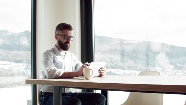 A mature man with glasses sitting at the table in new home, using tablet.