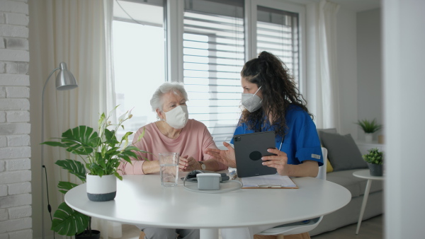 A healthcare worker or caregiver with tablet visiting senior woman indoors at home, explaining. Coronavirus concept.