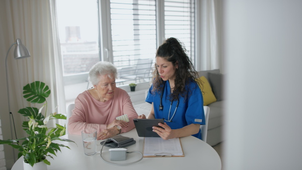 A healthcare worker or caregiver with tablet visiting senior woman indoors at home, explaining.