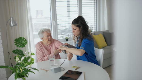 A healthcare worker or caregiver visiting senior woman indoors at home, measuring her blood pressure.