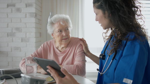 A healthcare worker or caregiver with tablet visiting senior woman indoors at home, explaining.