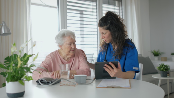 A healthcare worker or caregiver with tablet visiting senior woman indoors at home, explaining.