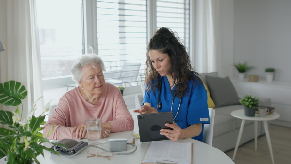 A healthcare worker or caregiver with tablet visiting senior woman indoors at home, explaining.