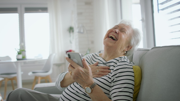 A senior woman at home with smartphone having an video call and laughing.