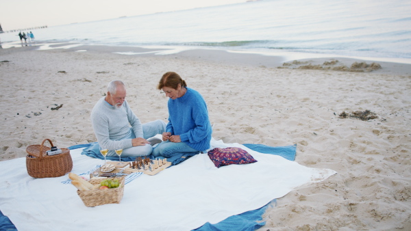 A happy senior couple in love outdoors on beach, having a picnic and playing chess.