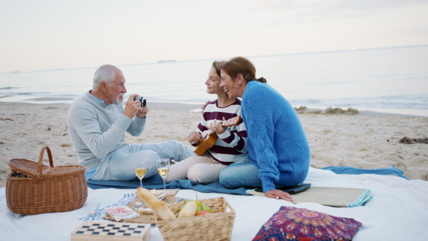 A happy senior couple with granddaughter on beach, having a picnic and taking pictures.