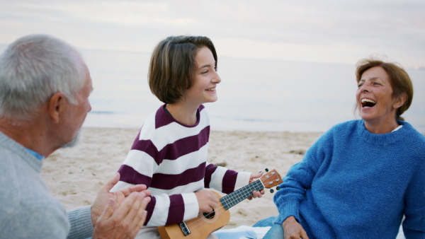 A happy senior couple with granddaughter on beach, having a picnic and playing guitar.