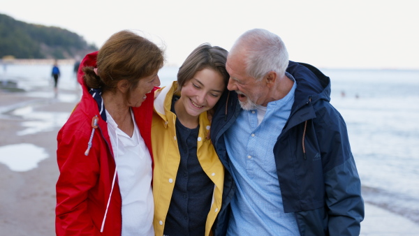 A happy senior couple with granddaughter on walk outdoors on beach at morning, hugging.